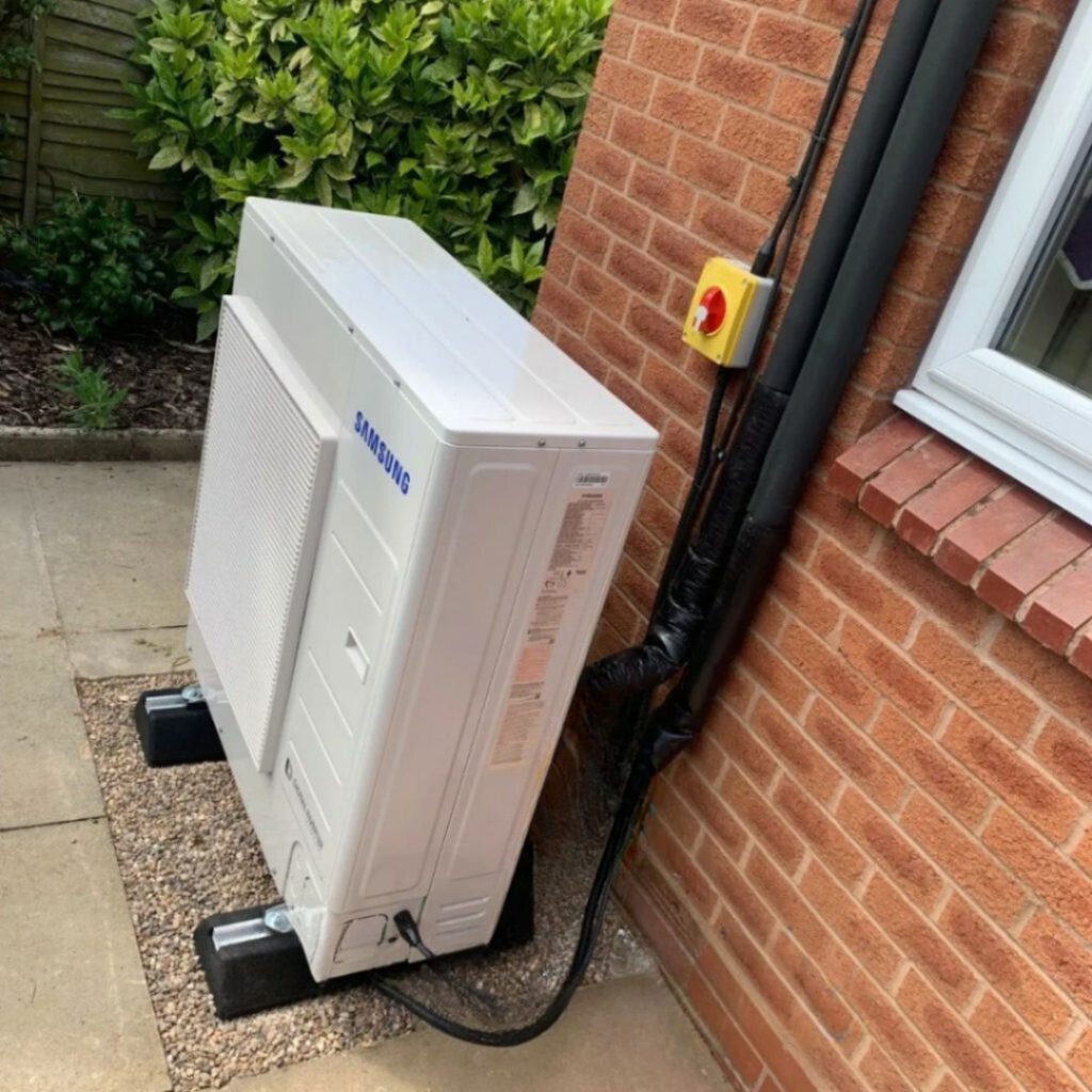 Outdoor air conditioning unit installed next to a red brick wall, with black tubing running along the wall and a green leafy background.